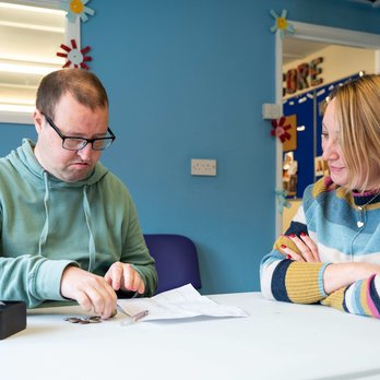 Matt is sitting on the left and has his budget page in front of him and his money tin by his hand. Claire sits next to him and is looking at his budget sheet, helping him make choices about what to put on his shopping list.