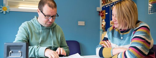 Matt is sitting on the left and has his budget page in front of him and his money tin by his hand. Claire sits next to him and is looking at his budget sheet, helping him make choices about what to put on his shopping list.