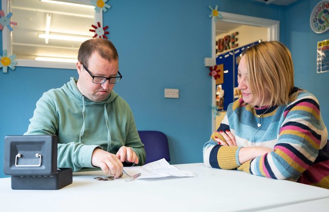 Matt is sitting on the left and has his budget page in front of him and his money tin by his hand. Claire sits next to him and is looking at his budget sheet, helping him make choices about what to put on his shopping list.