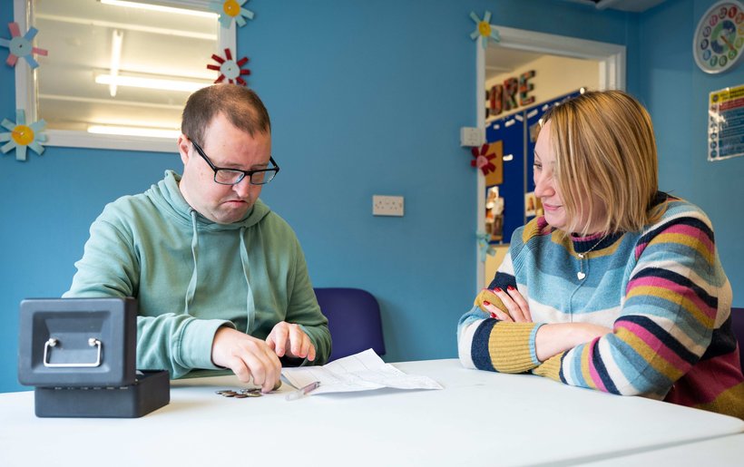 Matt is sitting on the left and has his budget page in front of him and his money tin by his hand. Claire sits next to him and is looking at his budget sheet, helping him make choices about what to put on his shopping list.