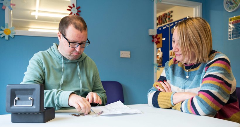 Matt is sitting on the left and has his budget page in front of him and his money tin by his hand. Claire sits next to him and is looking at his budget sheet, helping him make choices about what to put on his shopping list.