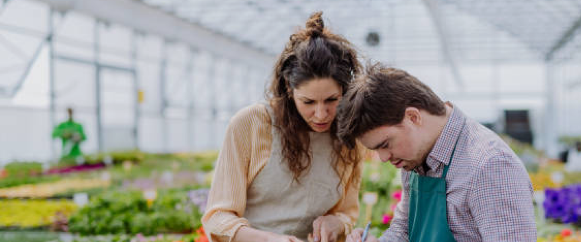 A young man with Downs is being supported in a horticultural environment. The young man is wearing a green apron, and is checking things off on a clip board, while his support worker is pointing to where he needs to write.
