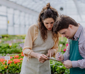 A young man with Downs is being supported in a horticultural environment. The young man is wearing a green apron, and is checking things off on a clip board, while his support worker is pointing to where he needs to write.