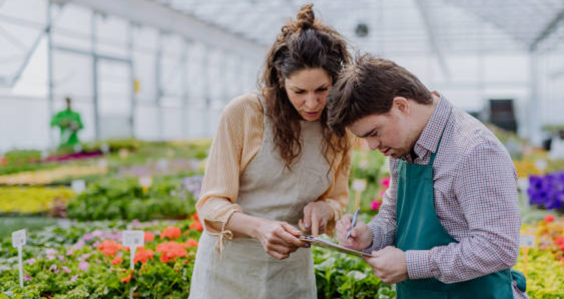 A young man with Downs is being supported in a horticultural environment. The young man is wearing a green apron, and is checking things off on a clip board, while his support worker is pointing to where he needs to write.
