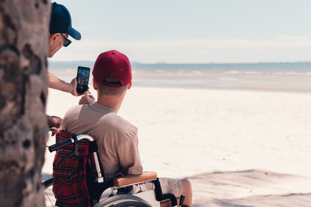 A young person in a wheelchair wearing a red cap has his back to us. He is looking out over the most glorious white sandy beach - the tide is out. Partially obscured by a tree trunk, his support worker is taking a selfie of them both with a smartphone.