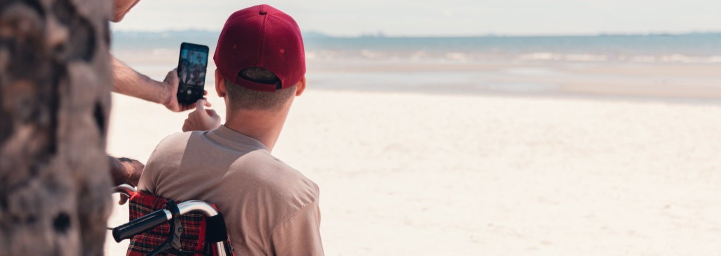 A young person in a wheelchair wearing a red cap has his back to us. He is looking out over the most glorious white sandy beach - the tide is out. Partially obscured by a tree trunk, his support worker is taking a selfie of them both with a smartphone.