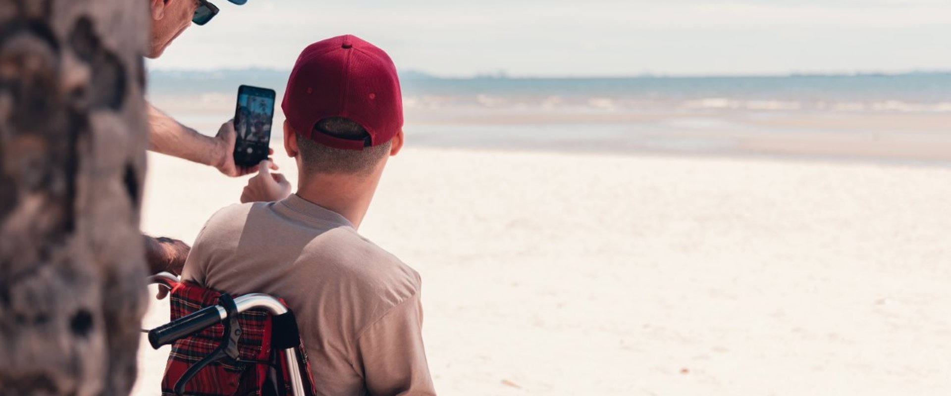 A young person in a wheelchair wearing a red cap has his back to us. He is looking out over the most glorious white sandy beach - the tide is out. Partially obscured by a tree trunk, his support worker is taking a selfie of them both with a smartphone.