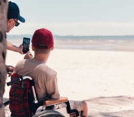 A young person in a wheelchair wearing a red cap has his back to us. He is looking out over the most glorious white sandy beach - the tide is out. Partially obscured by a tree trunk, his support worker is taking a selfie of them both with a smartphone.