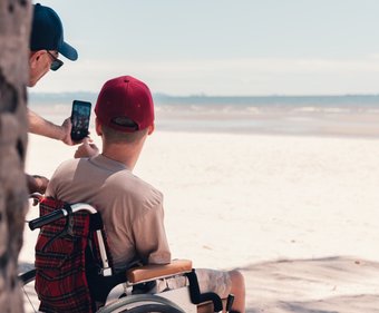 A young person in a wheelchair wearing a red cap has his back to us. He is looking out over the most glorious white sandy beach - the tide is out. Partially obscured by a tree trunk, his support worker is taking a selfie of them both with a smartphone.