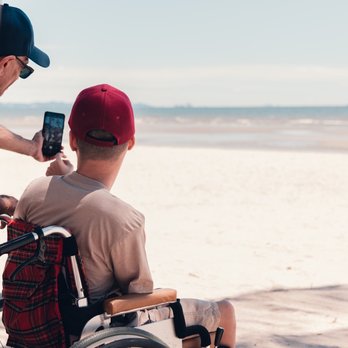A young person in a wheelchair wearing a red cap has his back to us. He is looking out over the most glorious white sandy beach - the tide is out. Partially obscured by a tree trunk, his support worker is taking a selfie of them both with a smartphone.