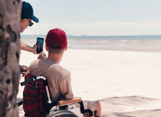 A young person in a wheelchair wearing a red cap has his back to us. He is looking out over the most glorious white sandy beach - the tide is out. Partially obscured by a tree trunk, his support worker is taking a selfie of them both with a smartphone.