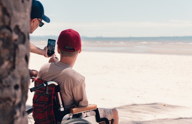 A young person in a wheelchair wearing a red cap has his back to us. He is looking out over the most glorious white sandy beach - the tide is out. Partially obscured by a tree trunk, his support worker is taking a selfie of them both with a smartphone.