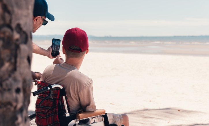 A young person in a wheelchair wearing a red cap has his back to us. He is looking out over the most glorious white sandy beach - the tide is out. Partially obscured by a tree trunk, his support worker is taking a selfie of them both with a smartphone.