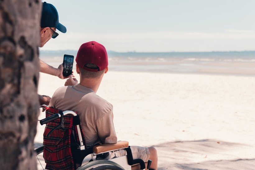 A young person in a wheelchair wearing a red cap has his back to us. He is looking out over the most glorious white sandy beach - the tide is out. Partially obscured by a tree trunk, his support worker is taking a selfie of them both with a smartphone.