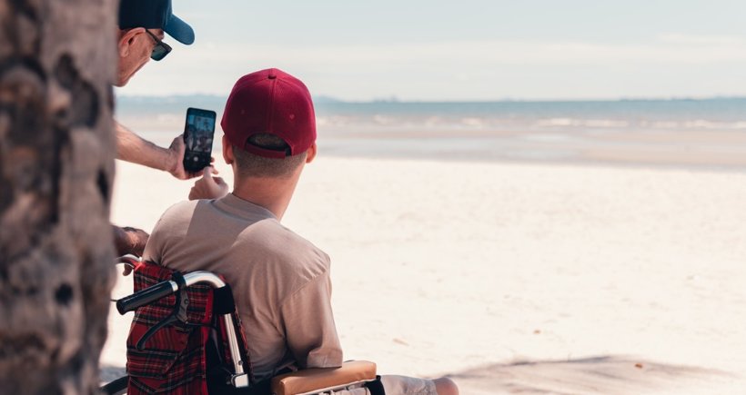 A young person in a wheelchair wearing a red cap has his back to us. He is looking out over the most glorious white sandy beach - the tide is out. Partially obscured by a tree trunk, his support worker is taking a selfie of them both with a smartphone.