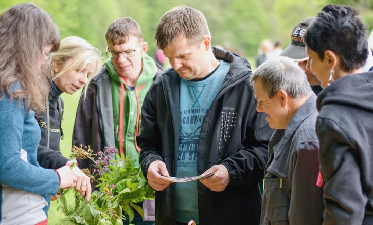 A group of four men are standing with their support worker, looking down at some plants. To the left, two women are pointing at the plants, telling the group something interesting about them.