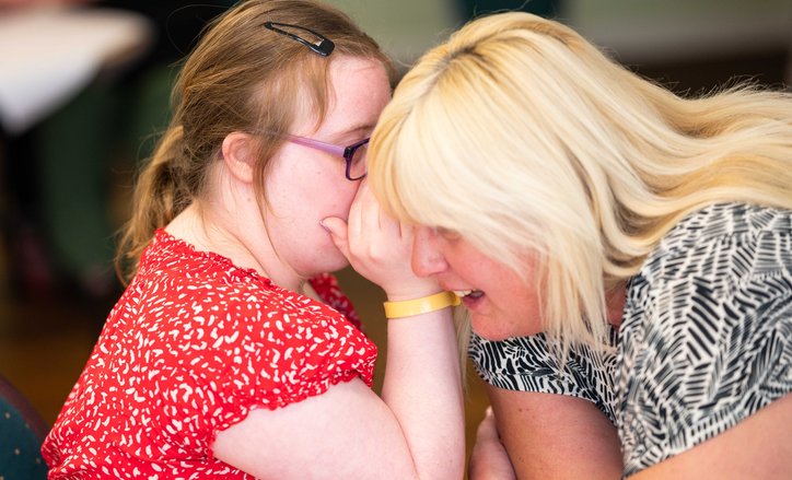 Emily, on the left, is wearing a beautiful bright red patterned top with frilly cap sleeves. You can only see a bit of her face though, as she has cupped her hand around her mouth while she whispers in her support worker's ear.