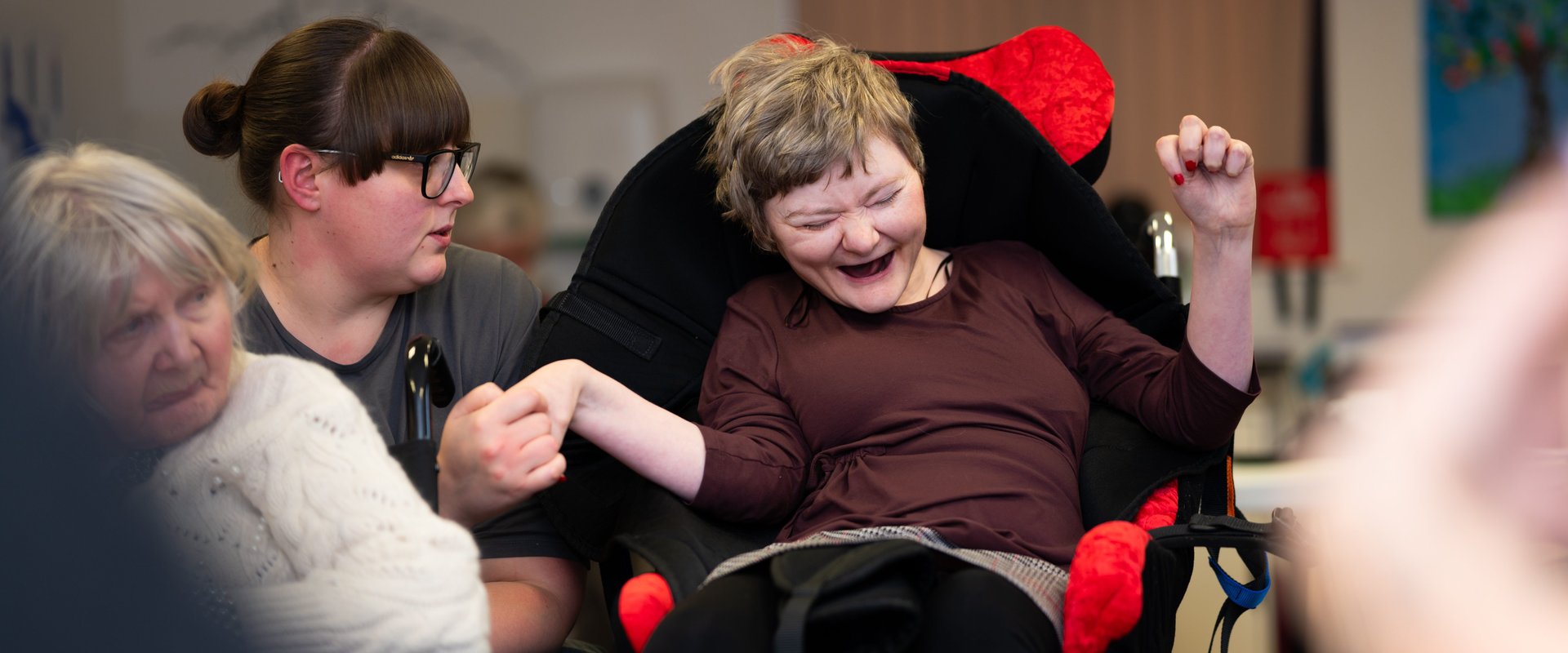 A person with complex disabilities holds her support worker's hand as she giggles. Her eyes are closed as she enjoys the moment. The support worker is looking at her, sharing her moment of joy.