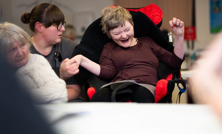 A person with complex disabilities holds her support worker's hand as she giggles. Her eyes are closed as she enjoys the moment. The support worker is looking at her, sharing her moment of joy.