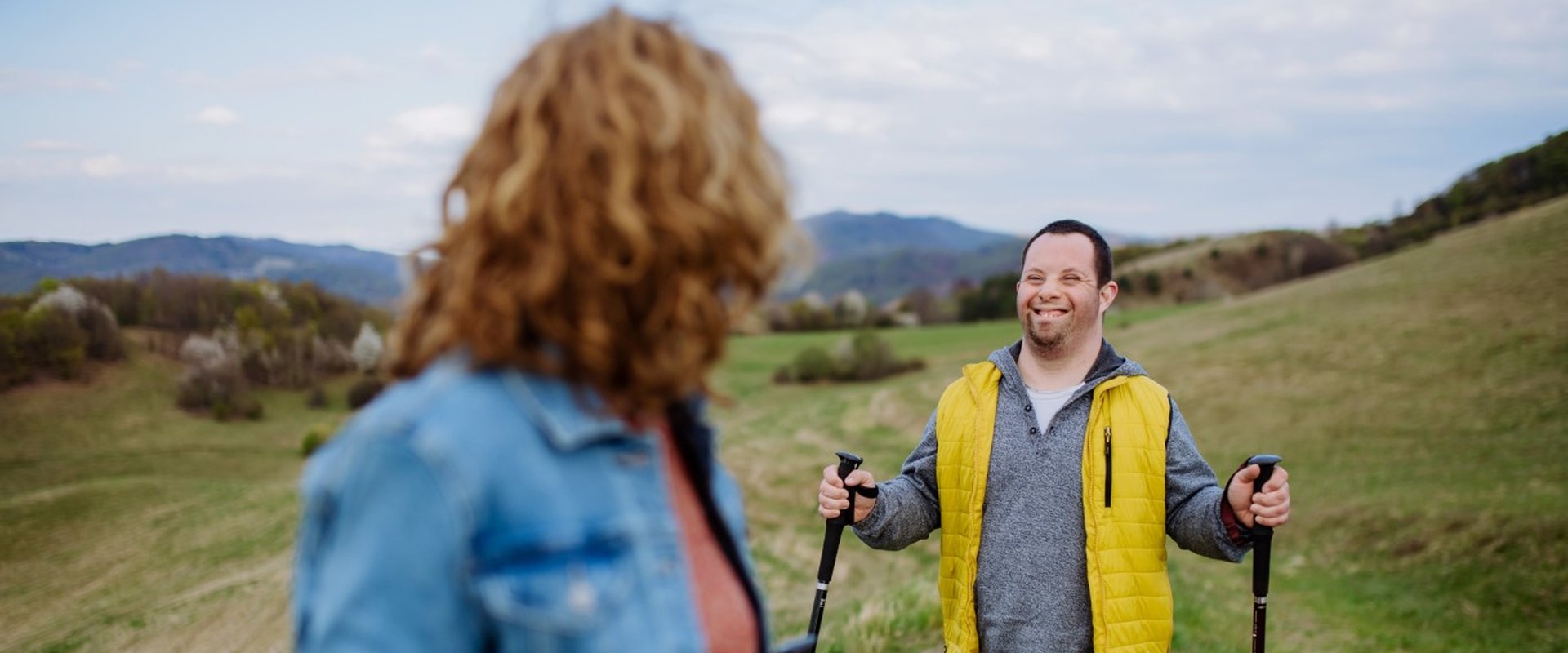 A young man with learning disabilities is enjoying walking in the countryside. There's a lovely view, with a hill in the distance behind him. He is wrapped up warm in a fleece and a bright yellow gilet, and he has Nordic walking poles.