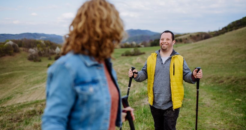 A young man with learning disabilities is enjoying walking in the countryside. There's a lovely view, with a hill in the distance behind him. He is wrapped up warm in a fleece and a bright yellow gilet, and he has Nordic walking poles.